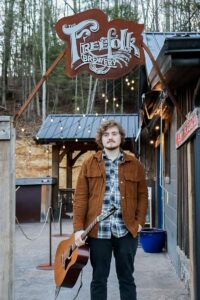 Gerald Potts, a musician, holds a guitar outside of a rustic, old building.