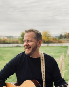 man in a field playing acoustic guitar.