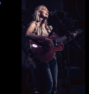 A woman sings and plays guitar on a dark stage. 